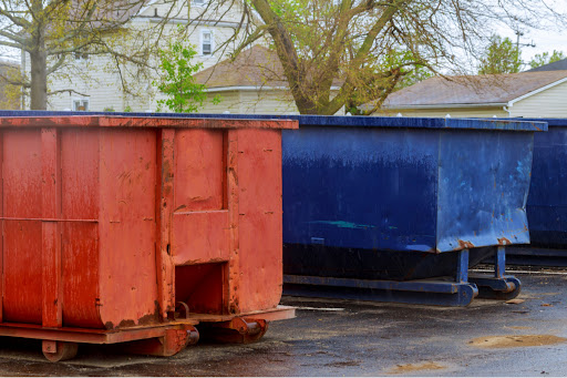 A red dumpster and blue dumpster sit in a parking lot waiting to be rented. There are homes in the background.