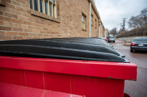 A red dumpster with black lid sits in a parking lot.