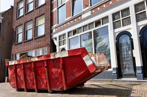 A red dumpster sits in the street in front of a group of homes and businesses.